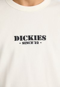 Biała, prążkowana koszulka z krótkim rękawem z haftowanym napisem "DICKIES - SINCE '22" w czerni z przodu. Okrągły dekolt i miękka faktura.