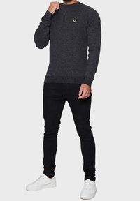 Pull tricoté gris charbon à col rond, avec un petit logo jaune sur le côté gauche de la poitrine, associé à un jean noir slim et des baskets blanches.