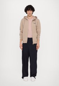 Hanorac bej cu fermoar și glugă cu șnur, tricou roz deschis și pantaloni largi bleumarin. Adidași albi completează ținuta.