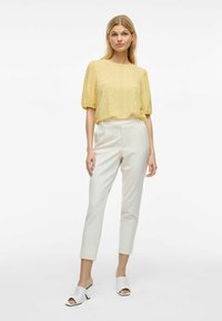 Blouse jaune à œillets avec manches bouffantes, associée à un pantalon blanc ajusté et des mules ouvertes blanches à accents noirs.