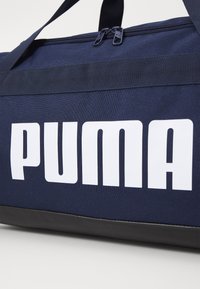 Sac de sport en tissu bleu marine avec un logo "PUMA" blanc. Forme rectangulaire avec deux fermetures éclair et un fond noir. Sangles d'épaule réglables.