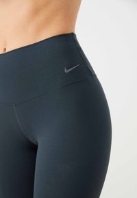 Wysokie, ciemnoniebieskie legginsy z gładką teksturą, z małym szarym logo Nike na pasie i płaskimi szwami dla komfortu.