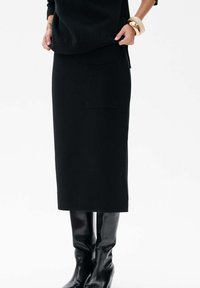 Kobieta w czarnej midi spódnicy z przednią kieszenią, czarnej długiej bluzce, czarnych wysokich butach do kolan oraz złotych i beżowych bransoletkach.
