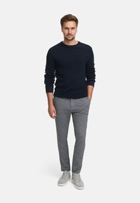 Marineblaue gerippte Pullover, hellgraue slim-fit Hosen und hellgraue Sneaker. Das Model steht mit den Händen in den Taschen vor einem schlichten Hintergrund.