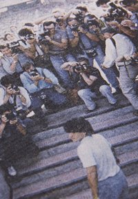 Een groep fotografen met camera's vul de kader en vangt een persoon in een wit shirt en jeans nabij een set stenen trappen.