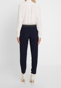 Navy byxor med en smal passform, elastisk midja och vit randdetalj. Bärs med en lätt, något genomskinlig blus och beige högklackade skor.