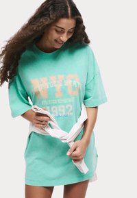 Myntgrön oversized t-shirt med NYC-grafik, kort ärm och avslappnad passform. Ett vitt band runt midjan ger kontrast. Texturerad bomullstyg.