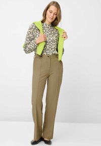 Blusa beige a fantasia con collo alto, pantaloni verde oliva con pieghe frontali, e un maglione verde brillante drappeggiato sulle spalle. Scarpe basse nere.