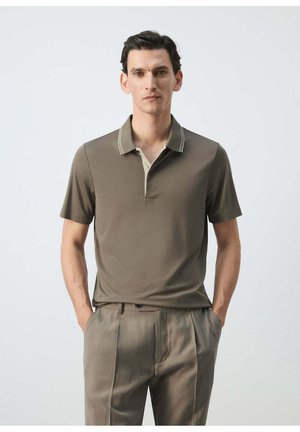 Hombre con polo de manga corta verde oliva y cuello en contraste y pantalones beige, de pie con las manos en los bolsillos sobre fondo liso.