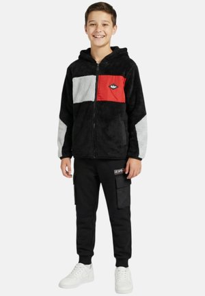 Garçon souriant portant une veste polaire noire zippée avec des panneaux gris et rouges, un pantalon cargo noir et des baskets blanches, debout sur un fond blanc.