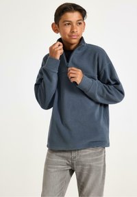 Blå fleecepullover med halvdragkedja och hög krage. Långa ärmar och en något avslappnad passform. Bärs med ljusgrå jeans.