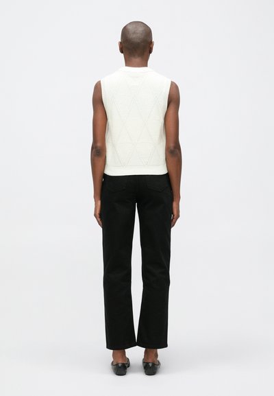 Top de punto sin mangas color crema con patrón de diamantes, combinado con pantalones negros de cintura alta y zapatos negros planos, visto desde la parte posterior.