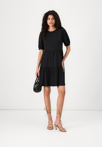 Vestido preto em camadas com mangas curtas bufantes, feito de um tecido suave. Usado com sandálias de tiras em tom bege e uma pequena bolsa preta. Design simples.