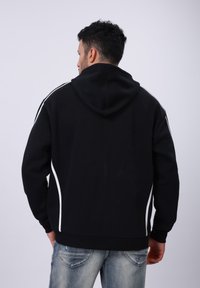 Zwarte hoodie met een grote capuchon, witte zijkanten met piping, ribbelboorden en een ribbelrand. Het materiaal lijkt zacht en gestructureerd. Draagt bij lichte blauwe jeans.