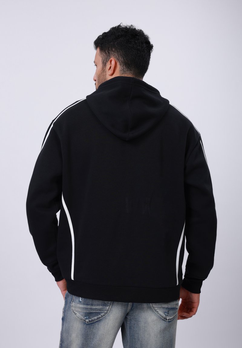 Felpa nera con cappuccio ampio, piping laterali bianchi, polsini e orlo a coste. Il materiale sembra morbido e dalla texture interessante. Indossata con jeans azzurri chiari.