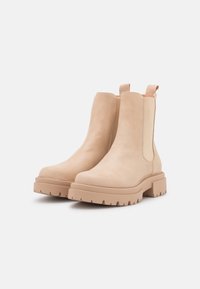 Botines de ante beige hasta el tobillo con paneles laterales elásticos, una suela de goma gruesa y estriada, y lengüetas para poner y quitar fácilmente. Textura suave y diseño limpio.