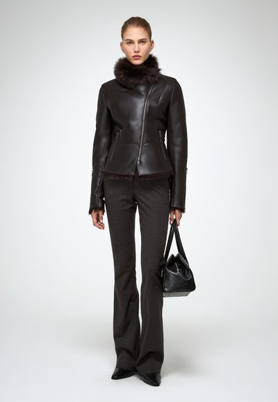 Donna con giacca di pelle nera con colletto di pelliccia, pantaloni neri su misura, scarpe a punta nere, che tiene in mano una borsa di pelle nera.