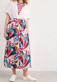 Chemise blanche à manches courtes et à boutons portée par-dessus un haut à rayures rouges et blanches, associée à une longue jupe à motifs vifs dans des tons de rose et de bleu.