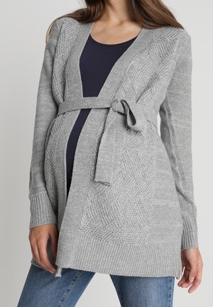 Personne enceinte portant un cardigan ceinturé en maille grise sur un haut foncé et un jean bleu, montrant un ventre de bébé.