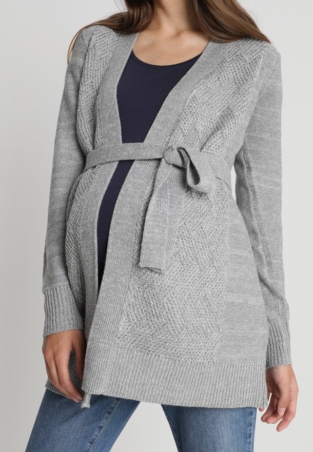 Personne enceinte portant un cardigan ceinturé en maille grise sur un haut foncé et un jean bleu, montrant un ventre de bébé.
