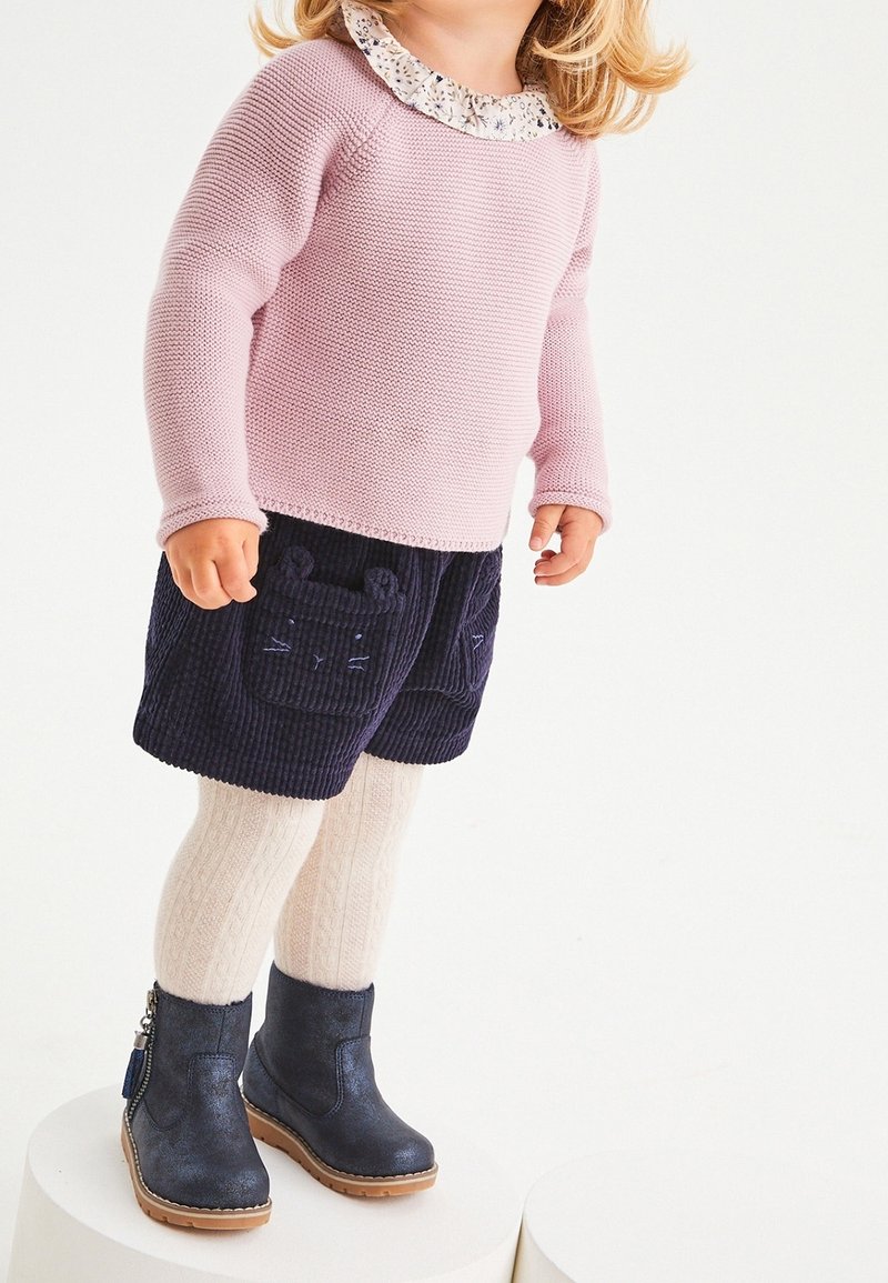 Un tout-petit portant un pull en maille rose, un short marine avec des visages d'ours, des collants crème et des bottes bleu foncé, debout sur des plateformes cylindriques blanches.