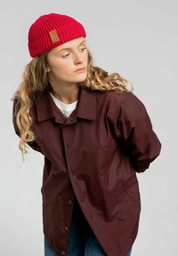 Bordowa wodoodporna kurtka z zatrzaskami i kieszenią, w zestawieniu z czerwoną dzianą czapką. Długie, faliste blond włosy i biała koszula pod spodem.