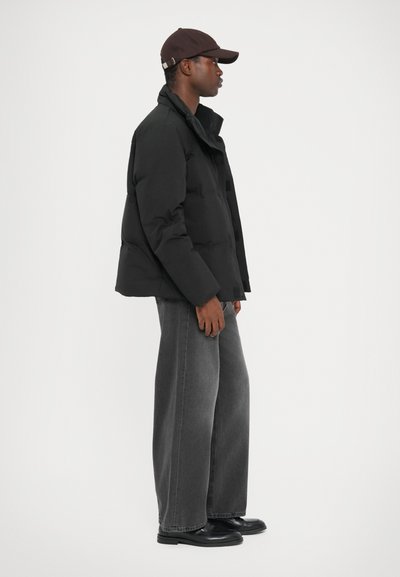 Black puffer jacket with a high collar, side pockets, and buttons, paired with wide-leg gray denim pants and black shoes. Brown cap on head.