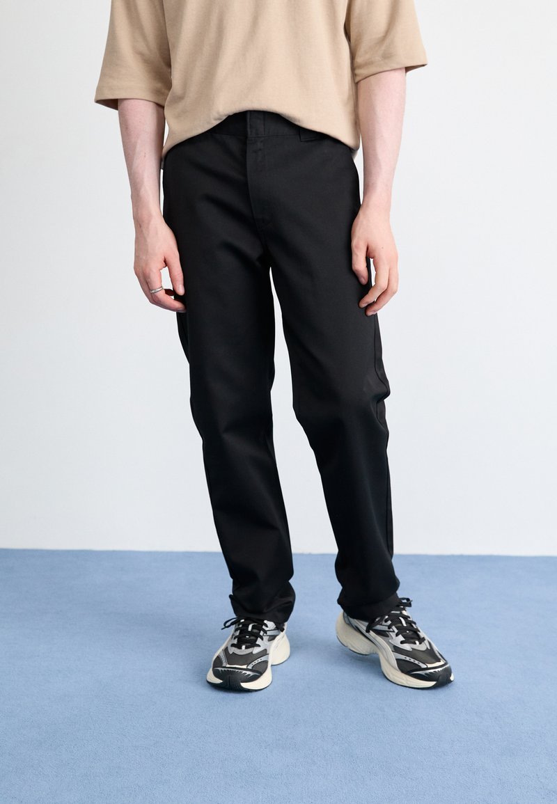 Sorte bukser med lige snit, parret med en beige oversized kortærmet skjorte og sort-hvide sneakers på en blå overflade.