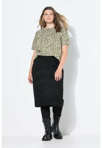 Grünes Paisley-Muster Kurzarmoberteil, kombiniert mit einem schwarzen strukturierten Bleistiftrock und schwarzen Stiefeletten. Das Model steht selbstbewusst vor einem weißen Hintergrund.