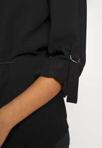 Chemise noire avec des manches roulées maintenues par des anneaux métalliques, présentant une taille cintrée et une texture lisse. Le tissu semble léger et flexible.