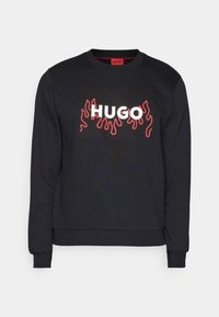 Sweat-shirt noir à manches longues avec motif flamme rouge et texte blanc "HUGO" centré sur le devant, doté de poignets et ourlet côtelés.