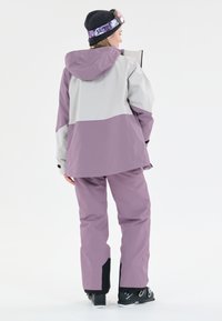 Lila och grå skidjacka med huva och matchande byxor, med en sleten struktur och kontrasterande svarta detaljer. Synliga skidstövlar.