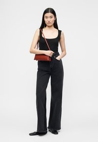 Γυναίκα με μακριά σκούρα μαλλιά που φοράει μαύρο τοπ χωρίς μανίκια και φαρδιά μαύρα τζιν, κρατώντας μια κόκκινη τσάντα ώμου, στέκεται σε λευκό φόντο.