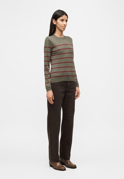 Πουλόβερ ελιάς  με οριζόντιες μπορντό ρίγες, λαιμόκοψη σε στρογγυλή γραμμή και μακριά μανίκια, σε συνδυασμό με παντελόνι σκούρου καφέ με ίσια γραμμή και καφέ μπότες με τακούνι.