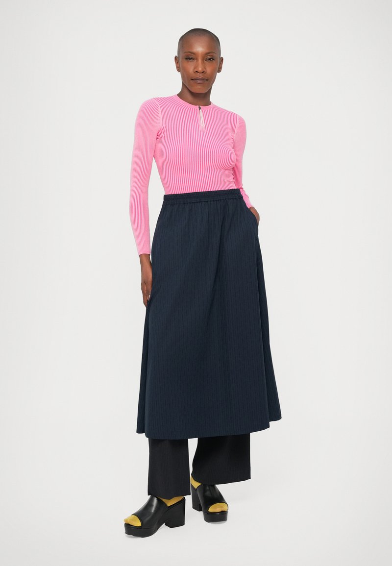 Pink ribbed long-sleeve top with a zip neck, paired with a flowy navy pleated skirt and chunky black clogs with yellow accents.