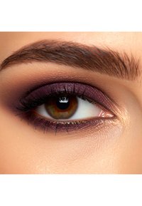 Close-up van een bruine oog met diepe paarse oogschaduw, lange zwarte wimpers en gedefinieerde wenkbrauwen tegen een lichte huidtint.