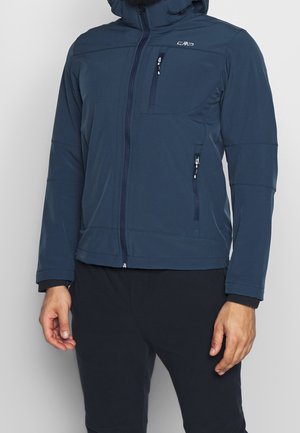Mann trägt eine marineblaue Softshell-Jacke mit Reißverschlusstaschen, Front-Reißverschluss und Kapuze, kombiniert mit schwarzen Hosen.