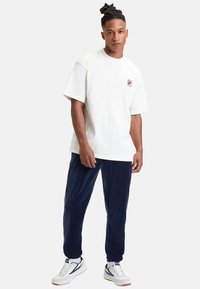 Biała bawełniana koszulka z krótkim rękawem z małym czerwonym i granatowym logo Fila na piersi, w połączeniu z granatowymi dresami z polaru i białymi trampkami.