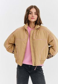 Mladá žena s hnedými vlasmi po pleciach má na sebe béžovú bundu z páperia cez ružový sveter a čierne džínsy, stojí so založenými rukami na páse.