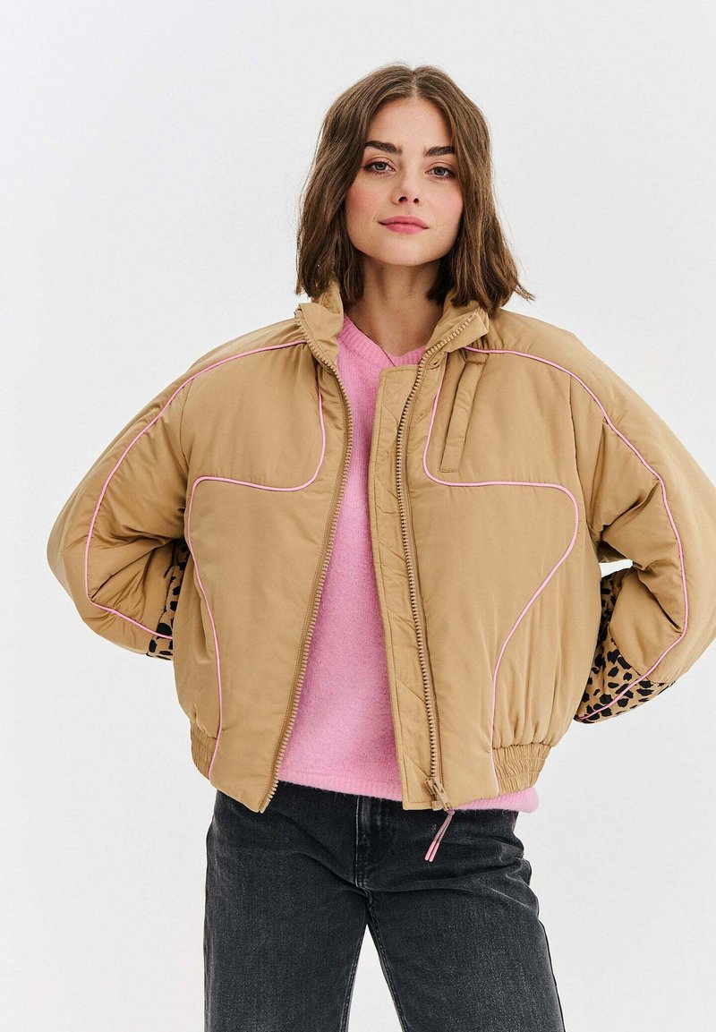 Mladá žena s hnedými vlasmi po pleciach má na sebe béžovú bundu z páperia cez ružový sveter a čierne džínsy, stojí so založenými rukami na páse.