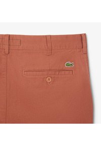Pantaloni corti di cotone in un colore arancio ruggine, caratterizzati da una sola tasca posteriore con chiusura a bottone e il logo del coccodrillo Lacoste ricamato in verde.