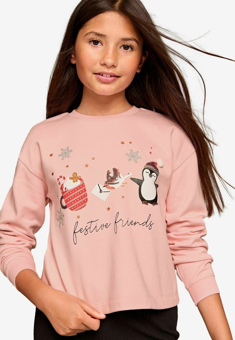 Sudadera rosa con un diseño gráfico festivo que incluye pingüinos, una taza y copos de nieve. El texto dice "amigos festivos". Tejido suave, mangas largas.