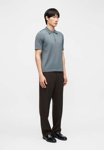 Άνδρας που φοράει μπλε πόλο με κοντά μανίκια, σκούρο καφέ παντελόνι και μαύρα επίσημα παπούτσια, στέκεται μπροστά σε ένα απλό λευκό φόντο.