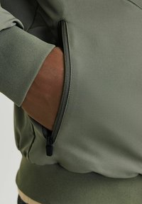 Sudadera verde con un tejido texturizado, un bolsillo con cremallera en el costado y puños acanalados, que muestra un diseño ajustado.