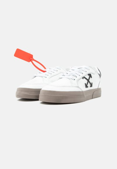 Λευκά sneakers χαμηλού τύπου με γκρι σόλες, μαύρο λογότυπο με σταυρωτό βέλος στα πλαϊνά, λευκά κορδόνια και φωτεινή πορτοκαλί ετικέτα με κορδόνι στη αριστερή παπούτσια.