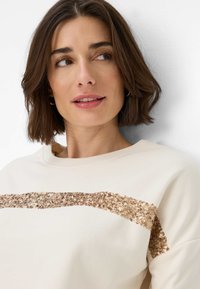 Cremegul sweatshirt med en tekstureret guldpalietstribe tværs over brystet. Har en rund halsudskæring og ribbede manchetter.