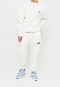 Felpa bianca e pantaloni jogger abbinati realizzati in tessuto morbido. La felpa presenta una piccola stampa di testo nera; i pantaloni jogger hanno un accento con logo verde. Sneakers blu chiaro.