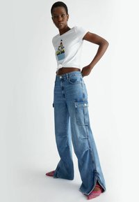 Camiseta blanca de algodón con corte corto y un gráfico colorido de cupcake, combinada con pantalones cargo de mezclilla azul claro de tiro alto y bolsillos laterales.
