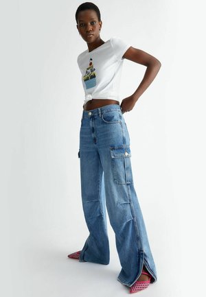 Camiseta blanca de algodón con corte corto y un gráfico colorido de cupcake, combinada con pantalones cargo de mezclilla azul claro de tiro alto y bolsillos laterales.