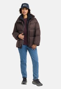 Doudoune marron avec un col haut, doublée en polaire grise, fermeture zippée, portée sur une chemise bordeaux, assortie avec un jean bleu et des baskets noires.